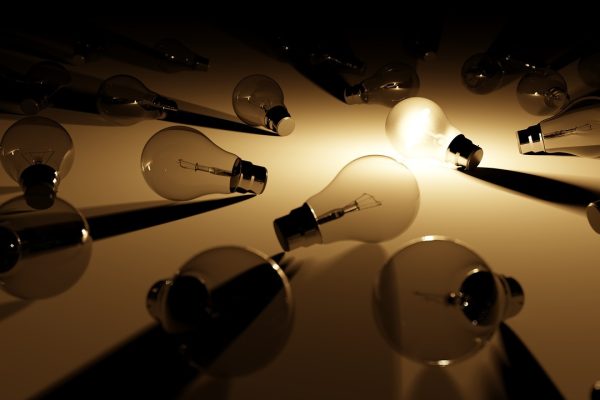 A single lightbulb is lit amidst a sea of unlit bulbs, all on their sides and facing different directions. The staging of the photo is dim, so the single bulb's illumination is made more prominent. It sits near the center and casts a white glow on the surrounding bulbs. Image via Pixabay.
