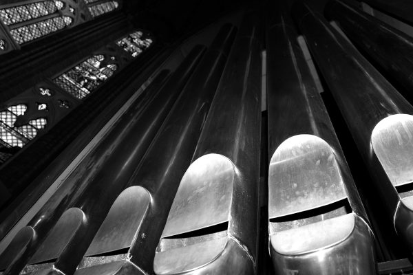 A black and white photo of a pipe organ in a church, taken from the perspective of a close-up shot at the base of the pipes. The pipes soar up towards the ceiling, giving the viewer the feeling of being small in the presence of something majestic. Tall stained-glass windows are visible to the left of the organ, although the scenes they depict are unclear. Image via Pixabay.