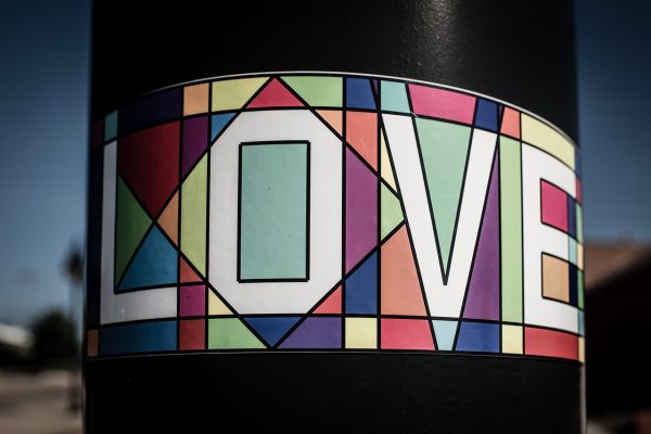A horizontal sticker with the word “Love” written in all-caps is adhered to a black pole. “Love” is written in white; the background of the sticker is a colorful rendition of a stained glass window, comprised of various shapes created by thick black lines. The background of the image is blurry, but it appears to be a sunny day; what looks like a palm tree can be seen on the left side of the image. Image via Pixabay.