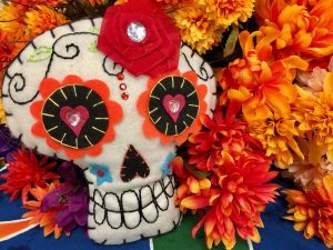 A cartoony-y felt skull made for the Day of the Dead is embroidered with hearts for eyes, flowers, and vine detailing. It sits amidst cloth flowers in rich hues of orange, yellow, red, and purple. The display sits on a colorful tablecloth made up of dark blue, green, white, and bright orange shapes. Image via Pixabay.