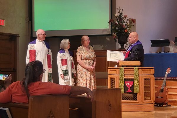 Rev. Richie Sanchez, Regional Minister of the Pacific Southwest Region of the Christian Church (Disciples of Christ) stands behind the pulpit of FCC Pomona and presents a certificate in celebration of the church’s 140th anniversary to (from left to right), Rev. Mike Fronk, Rev. Dr. Julie Roberts-Fronk, and Chris Abbott. Richie, Julie, and Mike are all clad in ministerial robes and stoles; Richie’s robe is black, and Mike and Julie’s robes are white. Mike and Julie are both wearing stoles handcrafted by FCC member Donna Graham that celebrate the church’s 140 year history; Richie’s stole is purple and red. Chris Abbott is wearing a long short-sleeved floral dress. All four people are angled towards one another and are situated at the front of the sanctuary. Other church members are visible from behind in the pews. Mike, Julie, and Chris are all middle-aged White people; Richie is a middle-aged Hispanic man.