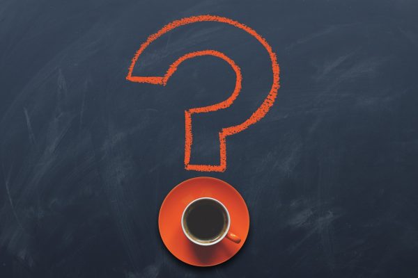 The outline of a question mark is drawn on a blackboard in orange chalk. The dot of the question mark is formed by a cup of coffee on an orange saucer.