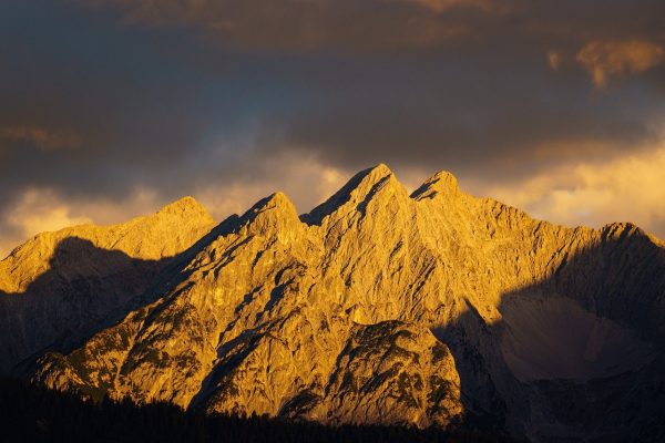 One of the craggy faces of a mountain range is illuminated in golden hues by the morning sun. The shadows of trees are visible along its base and along certain ridges; the mountain itself is also dappled with shadow. The blue sky in the background is partially obscured by the haze of dark clouds, although the clouds along the horizon are as radiant as the mountains. Image via Pixabay.