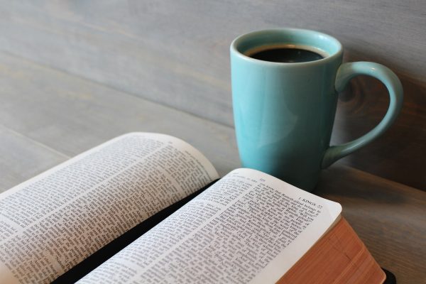 A Bible is open against a light gray table. A light turquoise mug filled with coffee, its handle turned to the right, sits above the Bible. A black ribbon bookmark runs down the center of the Bible; it is open to 1 Kings 22. Image via Pixabay.