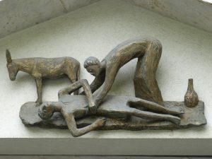 A sculptural interpretation of the story of the Good Samaritan. Said Good Samaritan is depicted kneeling over the man from Jericho, who grips his side in pain. A donkey stands in the background behind the man from Jericho, and a jug sits in the background behind the Good Samaritan. The sculpture appears to be affixed to a building near where the two sides of the roof meet. Image via Pixabay.
