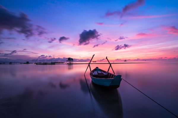 A simple wooden boat with two oars sits in still water at sunrise. It appears to be moored somewhere beyond the lower right corner. Other boats and marina infrastructure are visible in the distance. The sky and clouds are shades of pink, purple, and blue. Image via Pixabay.