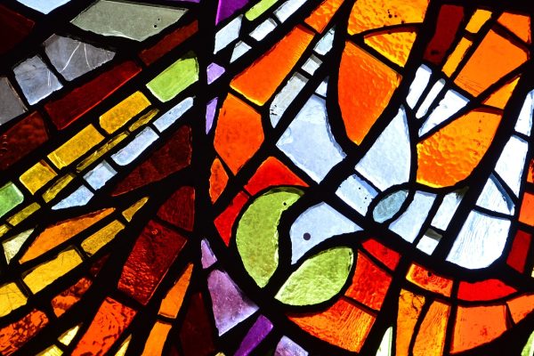 A stained-glass depiction of the Holy Spirit, shown as a dove descending with a green halo of glass around its head. The other panels of glass form a mosaic of vibrant orange, gold, burgundy, green, white, purple, and gray. Image via Pixabay.