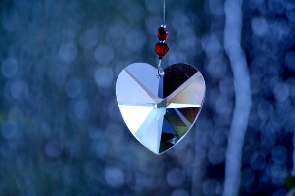 A prismed glass pendant in the shape of a heart hangs from a cord against a blurry blue background. Two heart-shaped red beads separated by two small clear beads sit on the cord above the heart. Image via Pixabay.