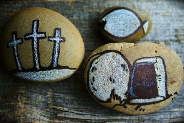 Three brown oval stones sit atop a warped wooden surface in a triangular formation. One point of the triangle depicts three crosses on a hillside drawn in black ink and white chalk. The next stone depicts the tomb with the stone rolled away, also drawn in black ink, white chalk, and blue chalk. The last point of the triangle is a smaller stone and depicts an ink and chalk rendition of a fish. Image via Pixabay.