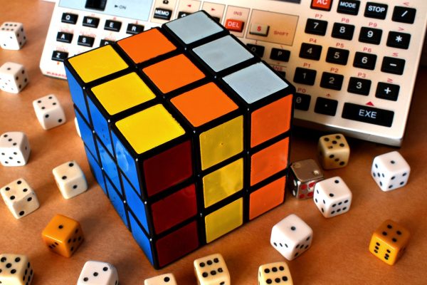 A partially solved red, yellow, orange, blue, and white Rubik’s cube sits amidst dice of various colors that have been scattered across a tabletop. The cube and the dice are in front of what appears to be some sort of calculator or adding machine. Image via Pixabay.