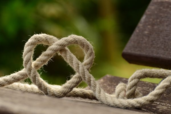A strand of rope is knotted to form the shape of a heart. It is sitting upright on a wooden surface; possibly a park bench or picnic table? The background is green but is not in focus. Image via Pixabay.