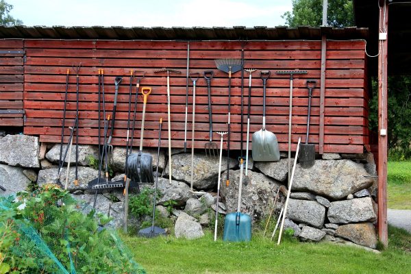 An array of yard tools—rakes, shovels, pitchforks, etc.—of various sizes are leaned against the wall of a wooden structure. Some are hanging from hooks on the same wall. The wooden structure sits on a base of several layers of large gray stones. The wooden and stone structure sits behind a plant covered in green netting and amid a yard of green grass. Image via Pixabay.