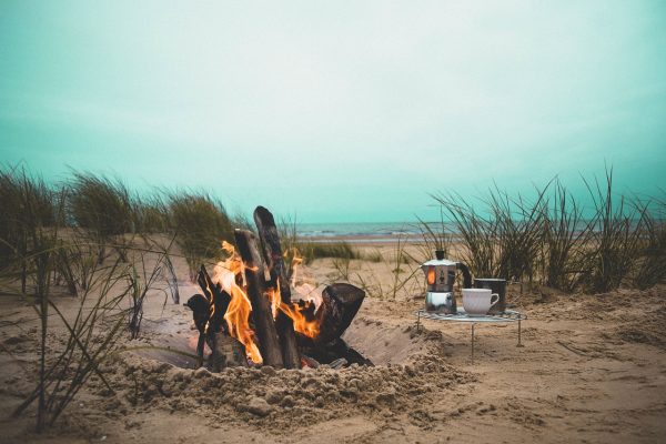A campfire burns on the beach in a sandy clearing lined with green reeds. A circular metal tray holding a coffee pot, white coffee cup, and large silver mug sits besides the fire. It is a cloudy day. The sky is a bright teal. The ocean and shore are visible in the background. Image via Pixabay.