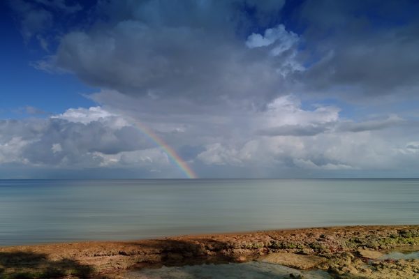 A photo of a rainbow stretching over the sea, as seen from the shore. A rocky stretch that looks like it forms tide pools is in the immediate foreground. It is a sunny day, and the sky is filled with puffy white clouds. The ocean is a pale green with streaks of blue. Image via Pixabay.