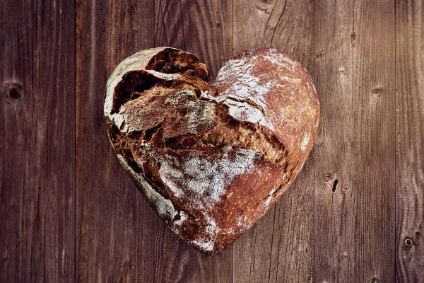 A heart-shaped loaf of bread sits atop a wooden surface comprised of horizontal brown boards. The loaf’s crust is cracked and dusted with flour. Image via Pixabay.