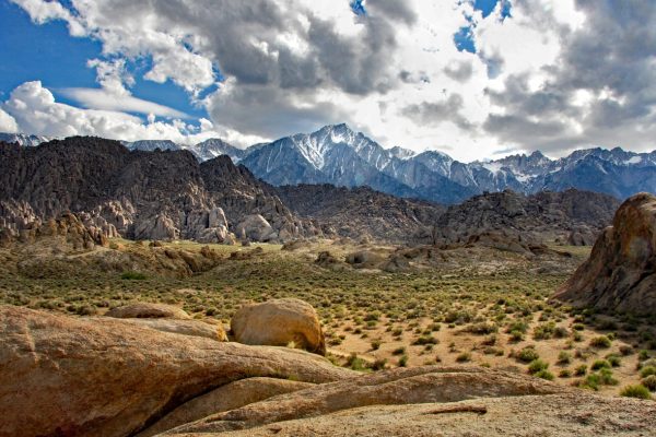 A photo of the Owens Valley in California. Tan rock formations interspersed with clumps of green brush give way to gray peaks, with snow-capped mountains along the horizon. It is a sunny day with puffy clouds occupying most of the bright blue sky. Image via Pixabay.