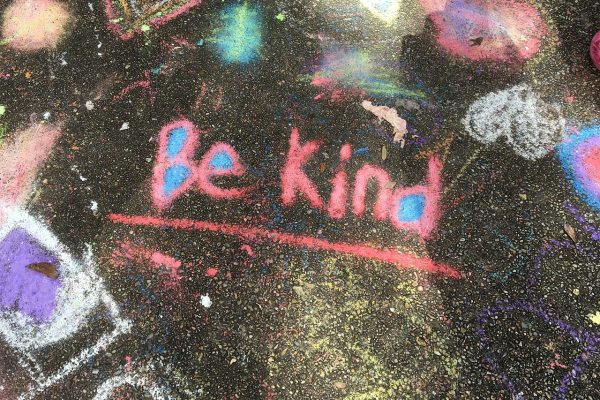 The words, “Be Kind” are written and underlined on asphalt in pink and blue chalk. Other shapes and squiggles in brightly colored chalk surround this central message. Image via Pixabay.