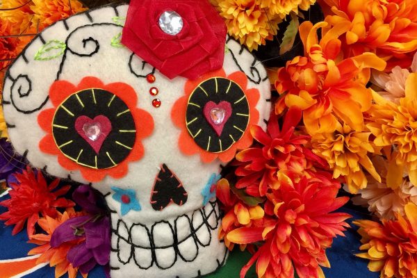 A felt skull is detailed with flowers, bold colors, and other whimsical stiches in the style of Día de los Muertos decorations. It is propped up against a backdrop of orange, gold, red, and purple silk flowers atop a blue, orange, green, and white tablecloth. The skull has a red flower pin made of gauzy fabric affixed to it like a bow and secured with a large clear rhinestone. The skull’s pupils are also rhinestones, albeit smaller, heart-shaped, and pale pink. Image via Pixabay.