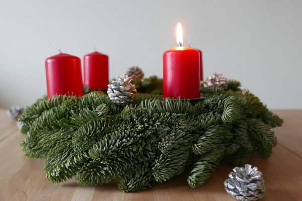An evergreen Advent wreath sits on a pale wooden table. The wreath has four short red candles arranged around its circumference, and the candle just right of center is lit. Silver pinecones sit around and atop the wreath.