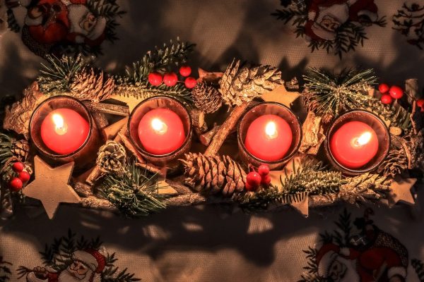 An Advent tableau as seen from above. All four red candles are lit in a rectangular display made of faux pine needles, holly, pinecones, and wooden stars. The tablecloth features embroidered Santa Clauses. Image via Pixabay.