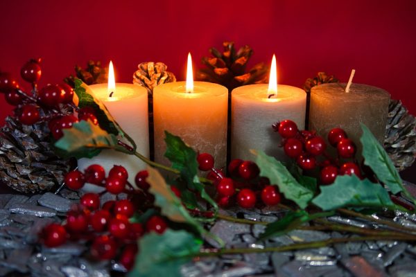 Four squat cylindrical candles are arranged in a horizontal row amidst sprigs of holly and pinecones. Three of the candles are lit. Image via Pixabay.