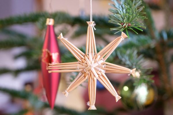 An ornament in the shape of a star hangs amidst evergreen sprigs. It is woven from thin strands of reed and has 8 points. Image via Pixabay.