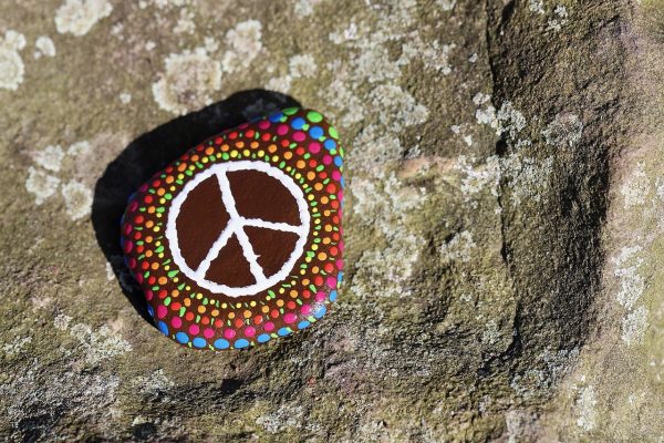 A rock painted with a white peace symbol. The peace symbol is surrounded by circles of brightly colored rainbow dots. Image via Pixabay.